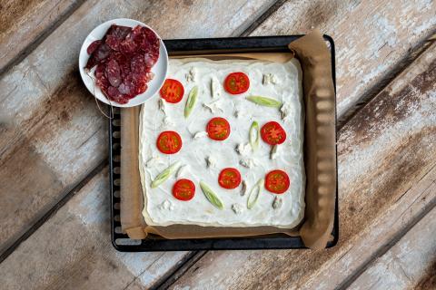 Mit Tomatenscheiben, Frühlingszwiebeln und etwas klein gezupftem Mozzarella belegen. Flammkuchen in den Ofen schieben. Nach ein paar Minuten herausnehmen mit der Wildschweinsalami belegen. (Quelle: Dorn/DJV)